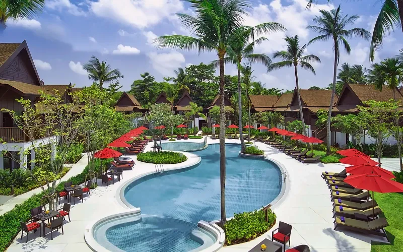 La piscina curva dell'Amari Koh Samui con ombrelloni rossi, lettini e palme da cocco davanti alle ali delle camere
