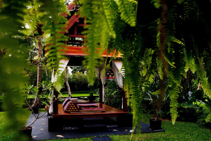 Anantara Bophut Koh Samuiの庭で、シダ越しに見える、クッション付きのタイ風の木造サーラー