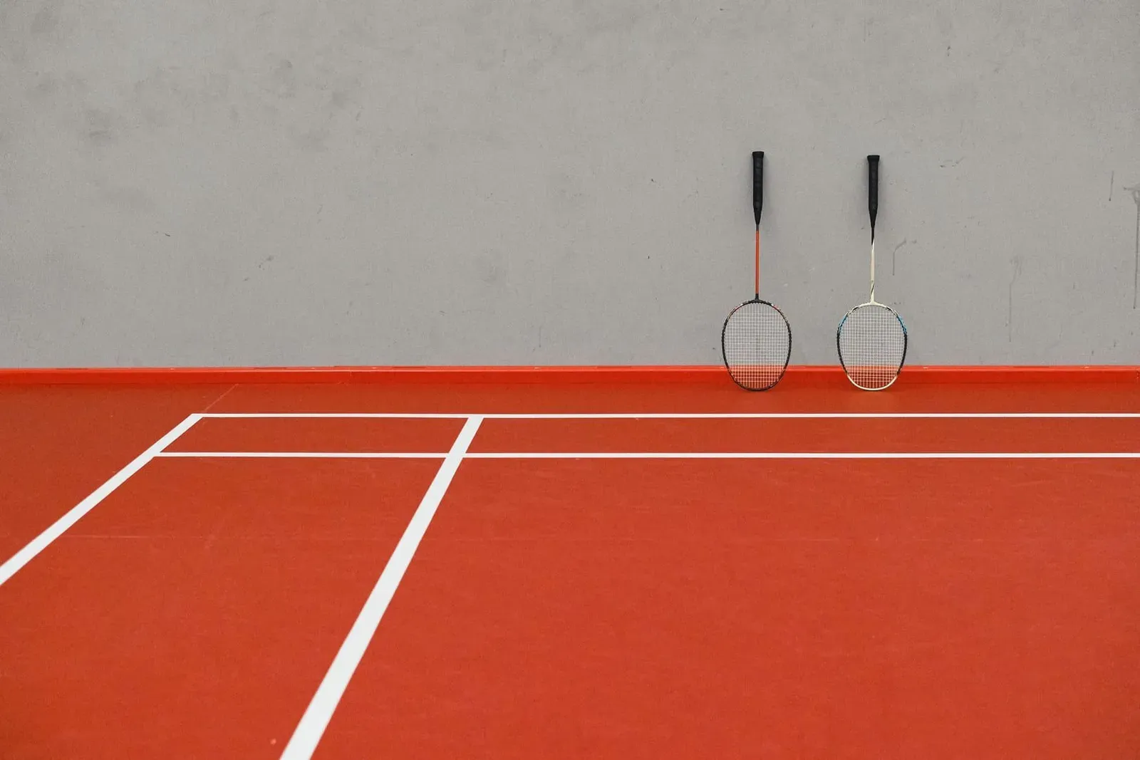 Two badminton rackets leaning against the wall of an empty red-floored indoor court