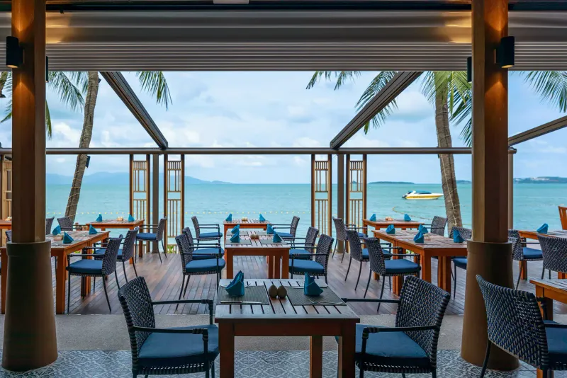 Terrazza del ristorante sulla spiaggia Chom Dao al Bandara Resort Samui con sedie di rattan e tovaglioli blu sulla baia