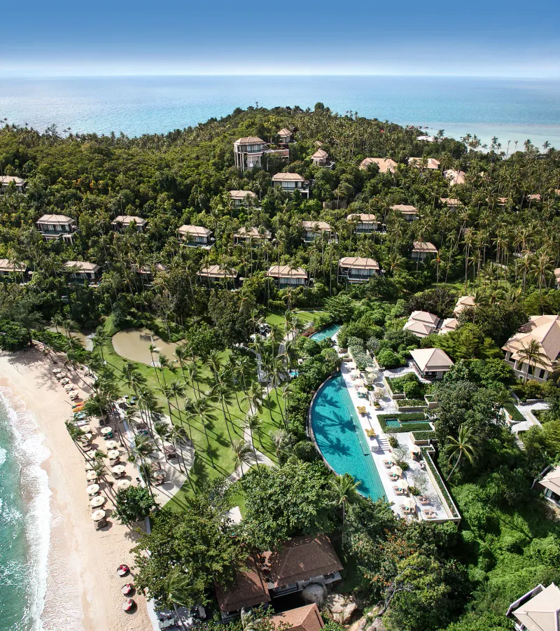 Veduta aerea del Banyan Tree Samui con la piscina curva, i prati, i lettini sulla spiaggia e le ville che salgono sulla collina