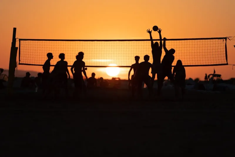 Silhouetted figures playing beach volleyball against a vivid orange sunset sky