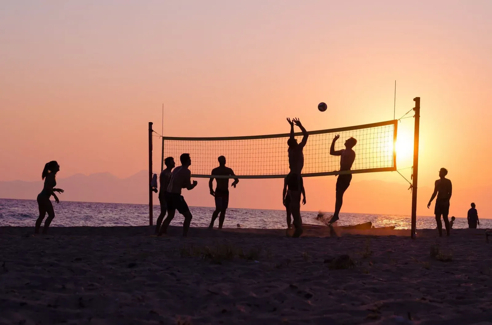 Silhouetted players set and spike a volleyball over a net on the beach as the sun sets over the water
