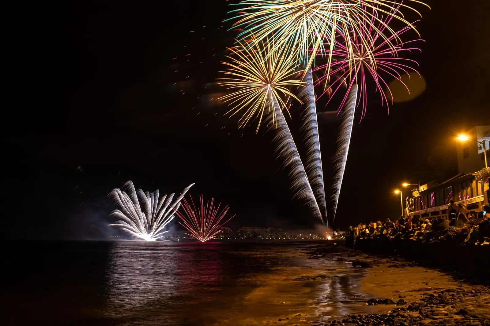 Fireworks reflecting on the ocean beside a crowded beach during a New Year countdown celebration