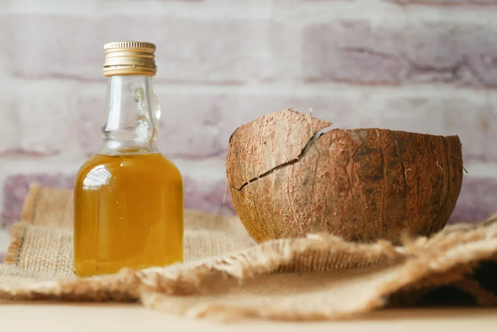 A small bottle of coconut oil beside a cracked open coconut shell on woven fabric