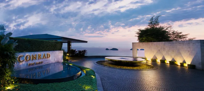 Entrée du Conrad Koh Samui au crépuscule avec enseigne éclairée, fontaine ronde et les Five Islands au fond