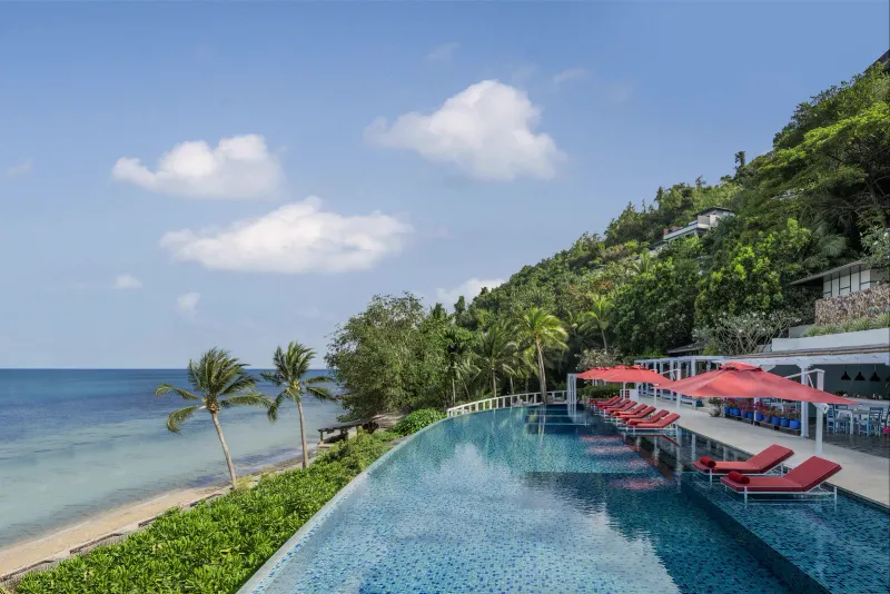 Piscine à débordement principale du Conrad Koh Samui avec transats rouges au bord de la baie et villas sur la colline