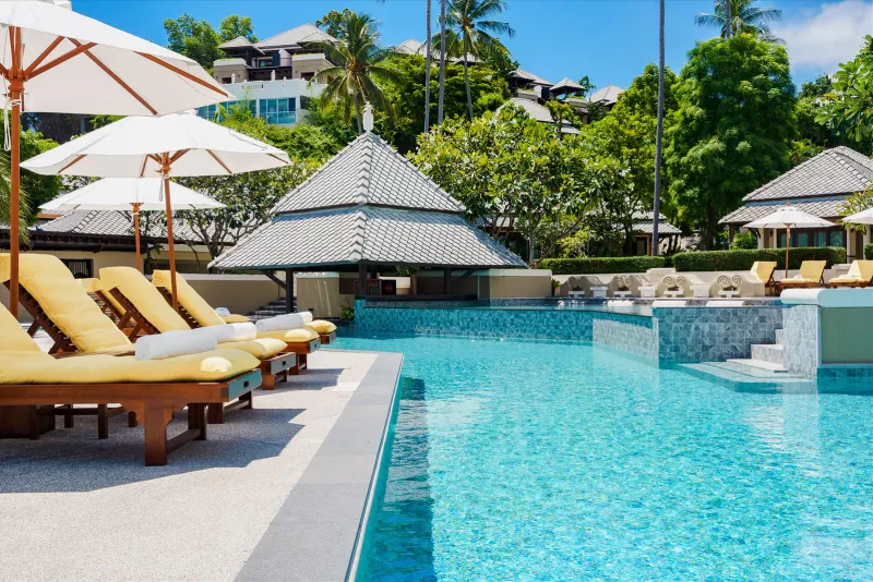 Piscine de Fair House Villas & Spa avec transats à coussins jaunes et pavillon du bar dans l'eau sous un versant de villas