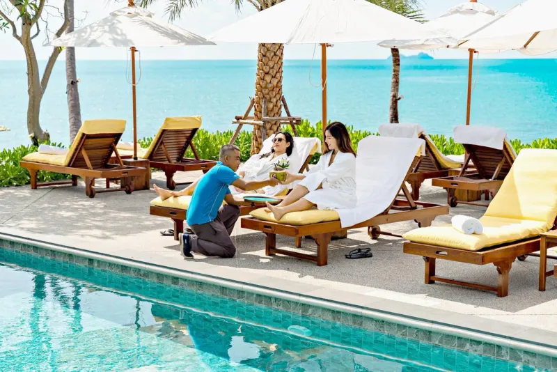 Employé servant une noix de coco fraîche à deux clients en peignoir sur des transats au bord de la piscine face à la mer