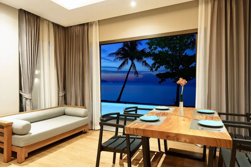 Salon et salle à manger d'une villa à la nuit tombée, avec une grande baie vitrée ouverte sur la piscine et la mer