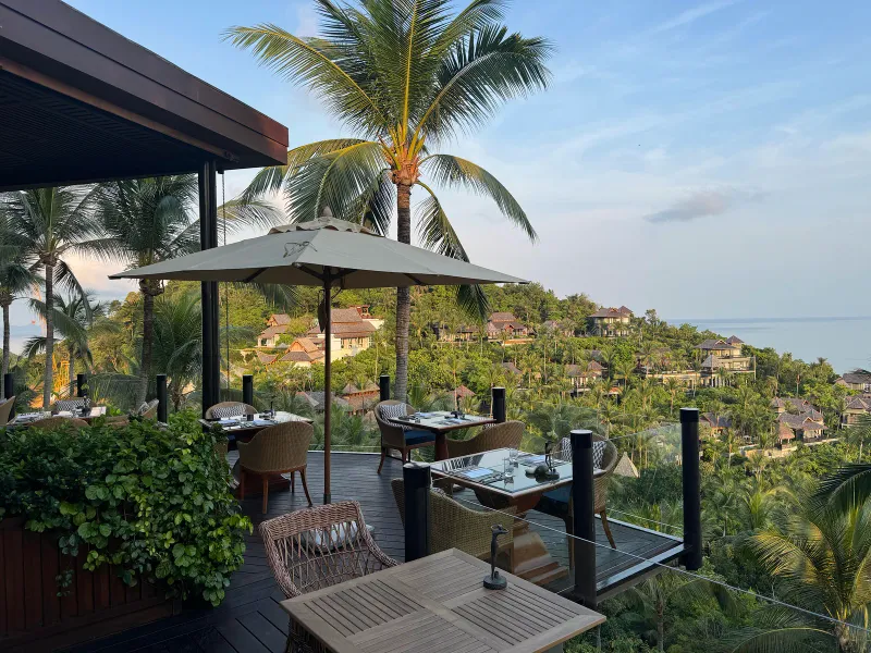 Terrasse de restaurant en plein air avec tables, parasol et palmiers au-dessus des villas de la colline au Four Seasons Resort Koh Samui