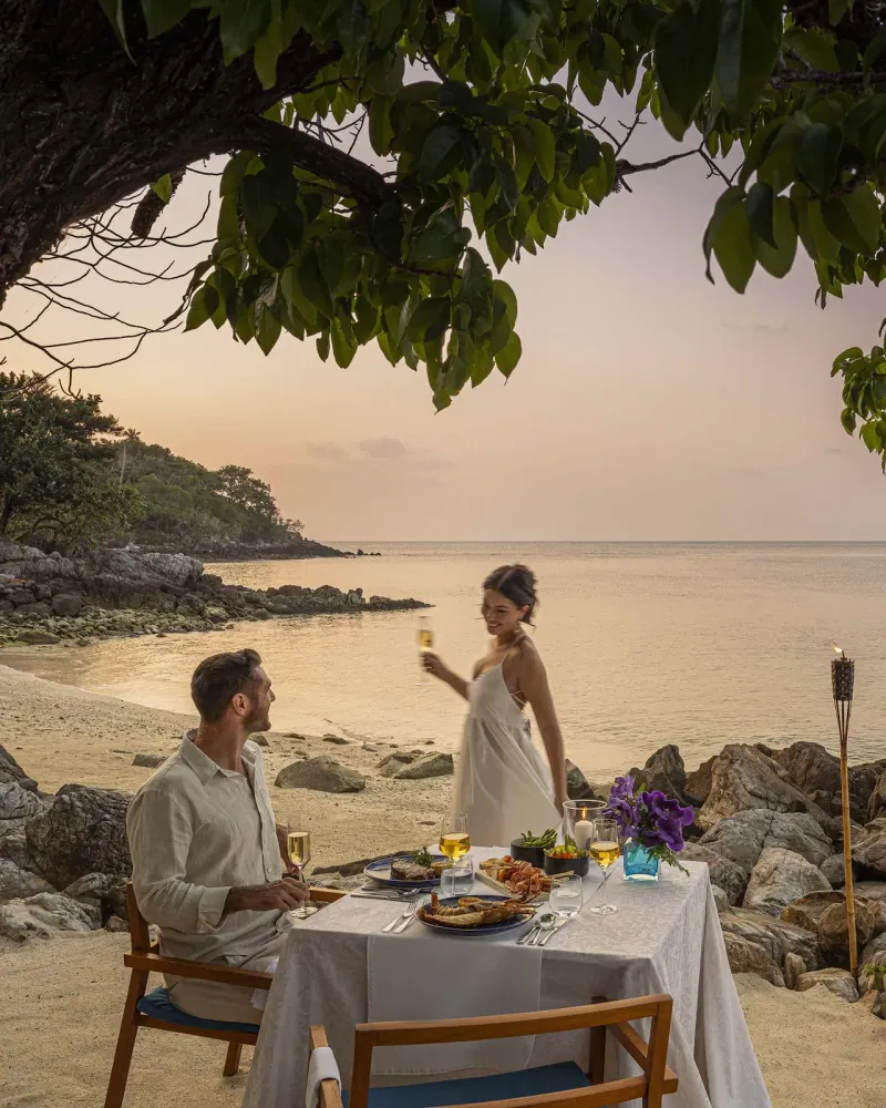 Couple à une table privée sur le sable à la tombée de la nuit, avec un rivage rocheux éclairé aux torches au Four Seasons Resort Koh Samui