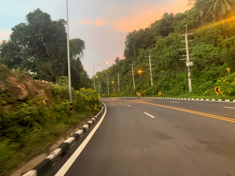 Rain-slicked hillside road with red rock cutting on the route toward Laem Yai Bay, Koh Samui