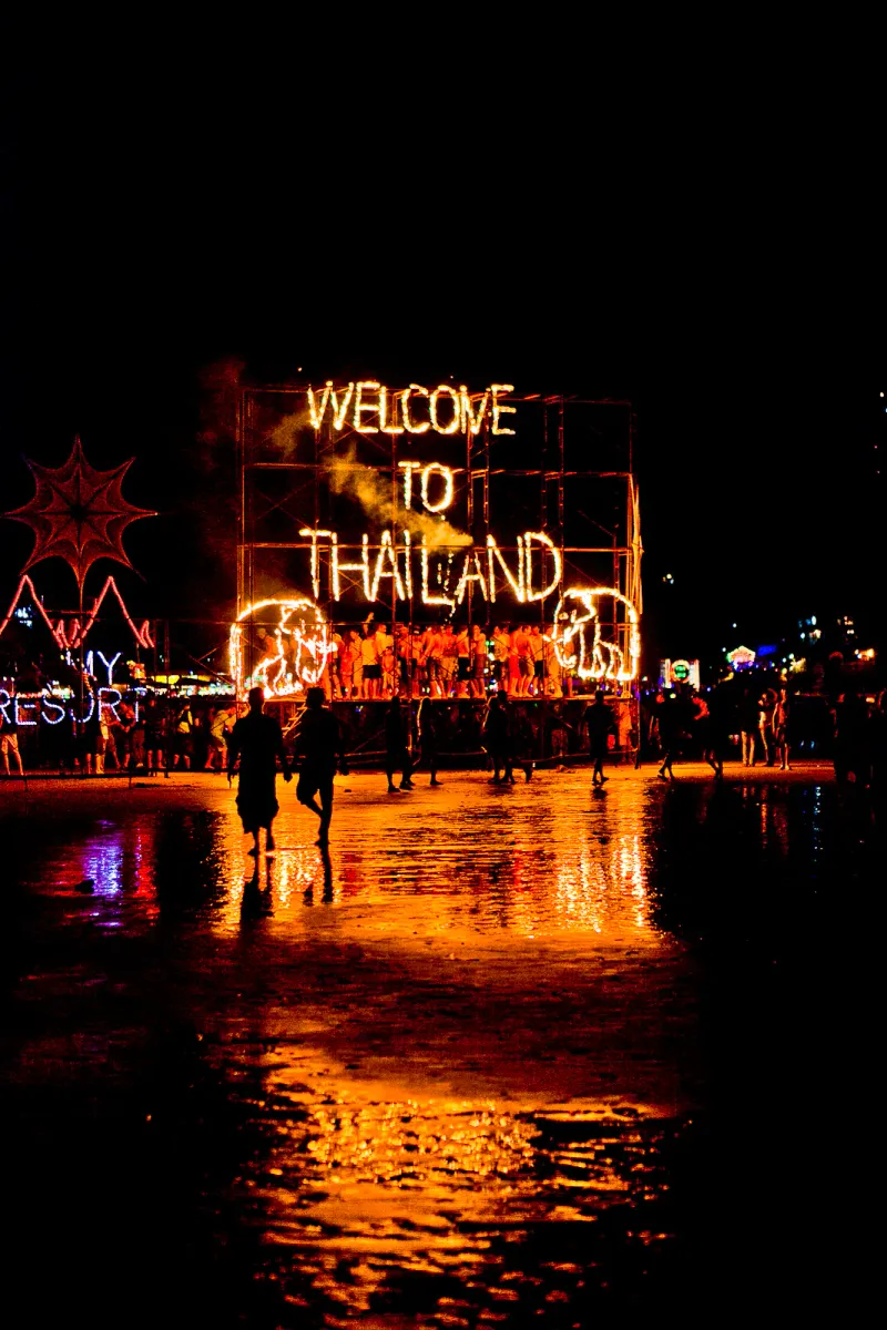 Night crowd on the beach at Haad Rin under a glowing Welcome to Thailand sign at a Full Moon Party on Koh Phangan