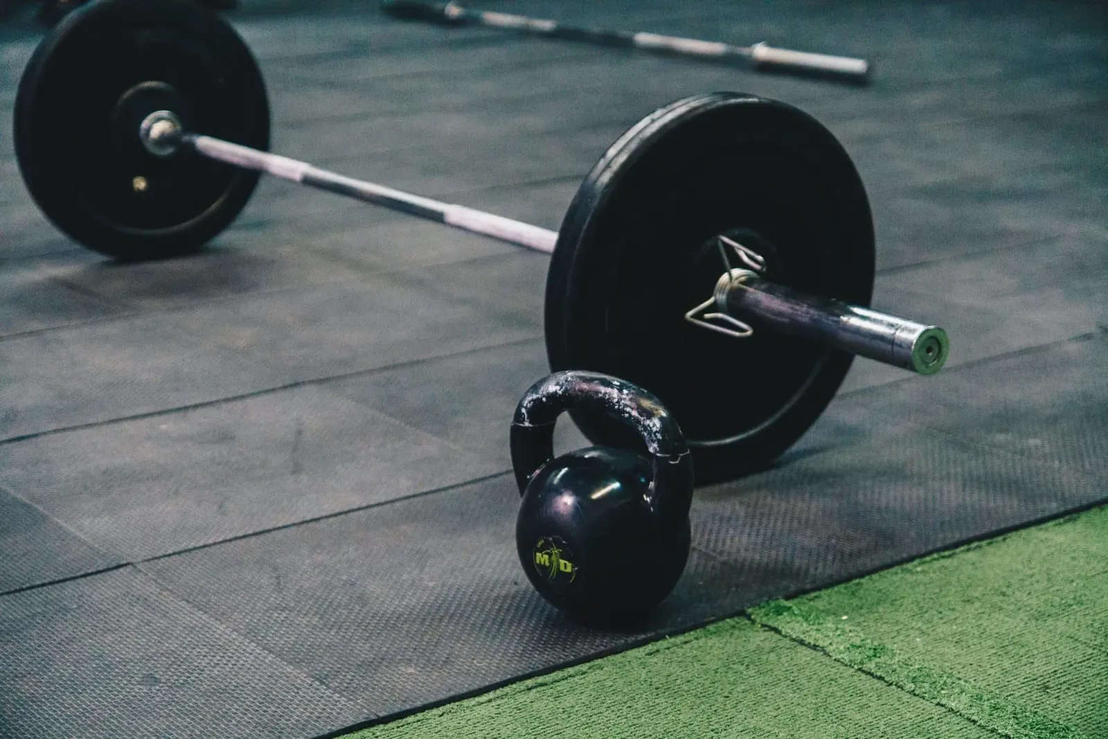 Loaded barbell and kettlebell resting on a rubber gym floor beside artificial turf strips
