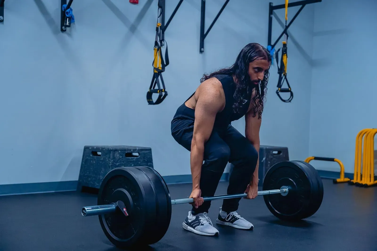 Man mid-deadlift lifting a loaded barbell off the floor in a gym with resistance bands hanging on the wall