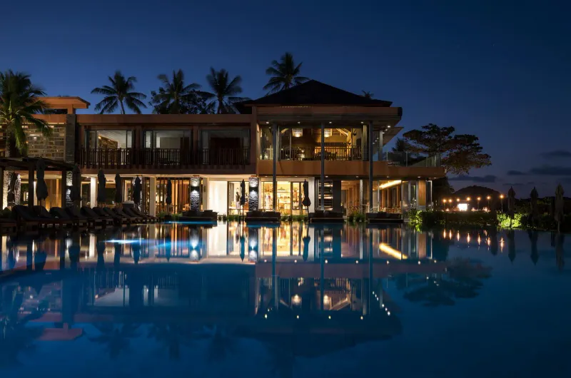L'Hansar Samui Resort al crepuscolo, con l'ala in terracotta riflessa nella piscina a sfioro, palme e terrazze illuminate