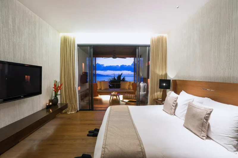 Camera da letto dell'Hansar Samui con un letto grande, televisore e uno scorcio di mare dalle porte del balcone