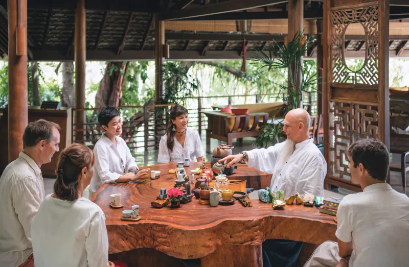 Ospiti e terapisti che condividono un tè attorno a un tavolo di legno a bordo grezzo in un padiglione aperto del Kamalaya Koh Samui