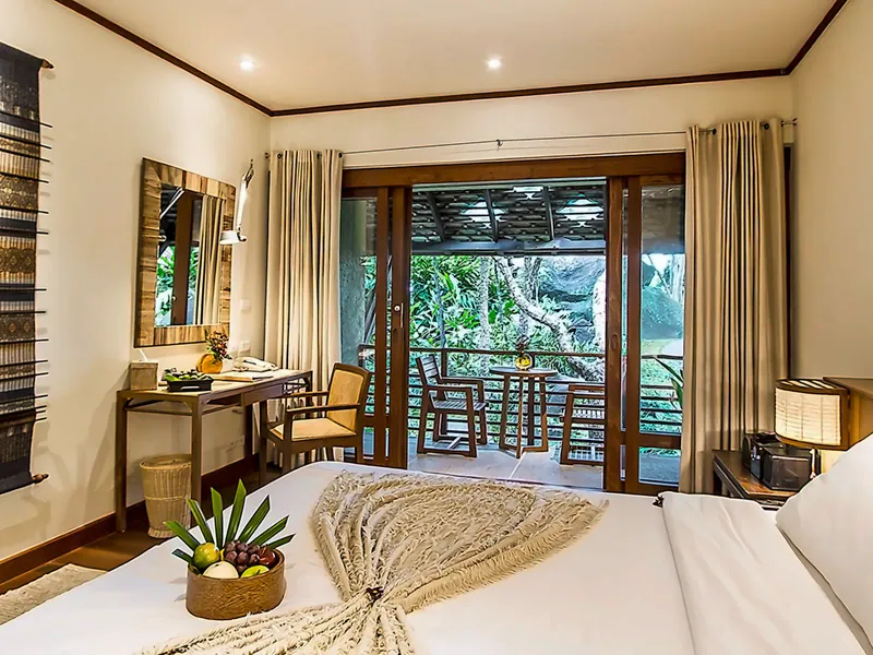 Camera da letto del Kamalaya Koh Samui con runner intrecciato sul letto, pavimento di teak e porte del balcone che si aprono sul giardino