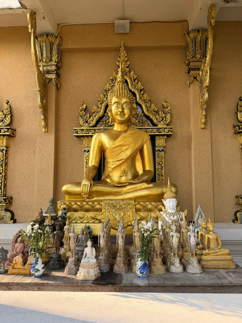 Large golden seated Buddha statue in a shrine alcove surrounded by rows of smaller votive statues and flowers