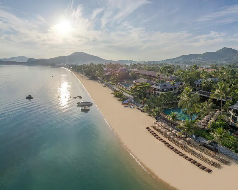 Vue aérienne d'une large baie de Koh Samui avec plage de sable, resorts parmi les palmiers et bateaux sur une eau calme