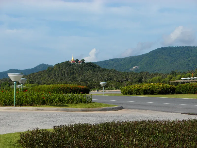 サムイ空港の外の道路と植栽の路肩。背後に森の丘が見える