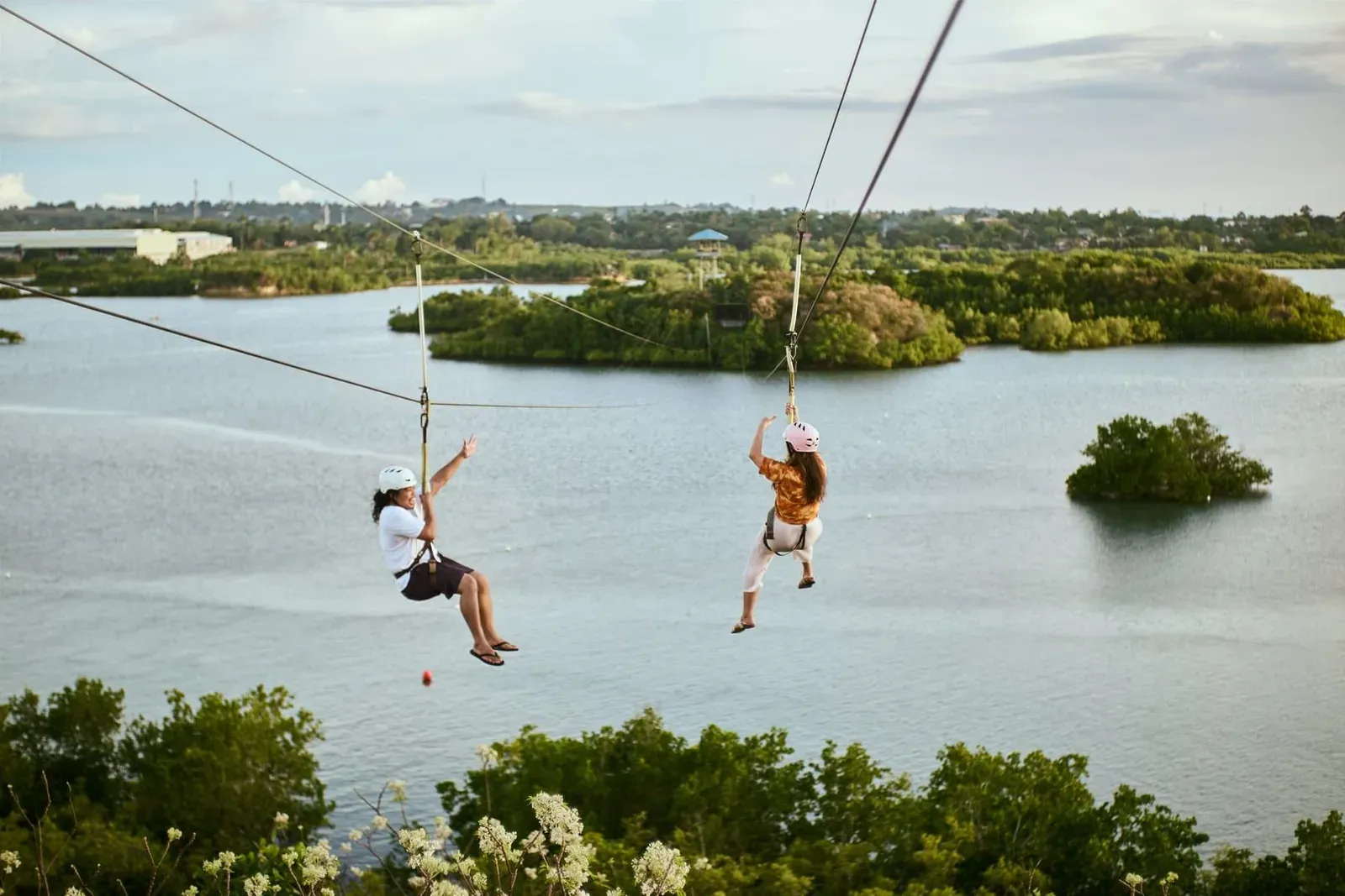 Two riders ziplining above calm water in a jungle adventure park similar to those on Koh Samui