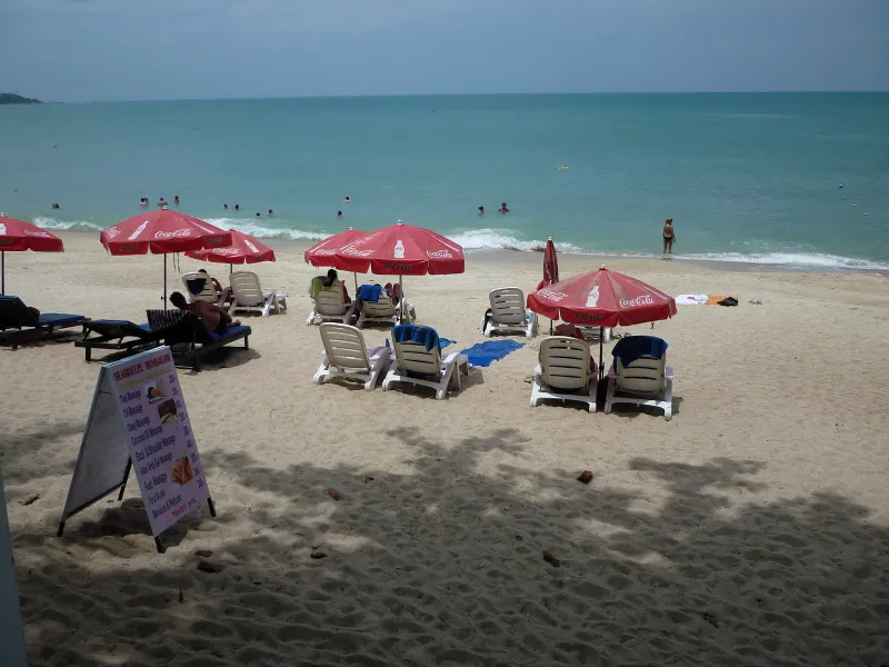 Red parasols and sun loungers on the sand at Lamai Beach, Koh Samui, with swimmers in the shallows