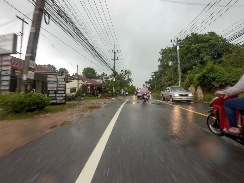 Route 4169 ring road through Lamai in heavy monsoon rain, shopfronts and a motorbike along the roadside