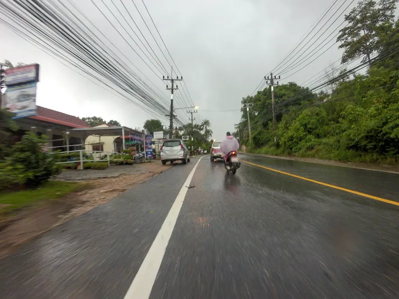 Cars and a motorbike on the wet ring road through Lamai village, Koh Samui, shops lining the roadside