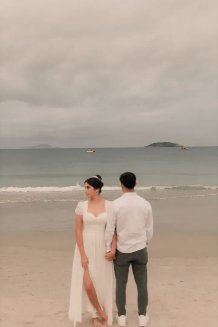 Couple in wedding attire holding hands on a quiet beach under cloudy skies