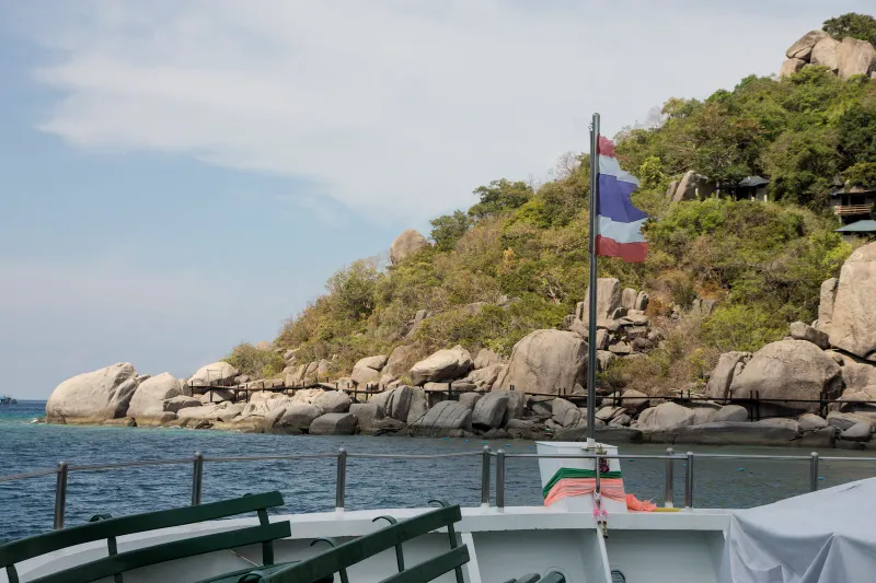 船のデッキから見た、タイの国旗と、岩がごろごろした木々の茂る島の岸辺
