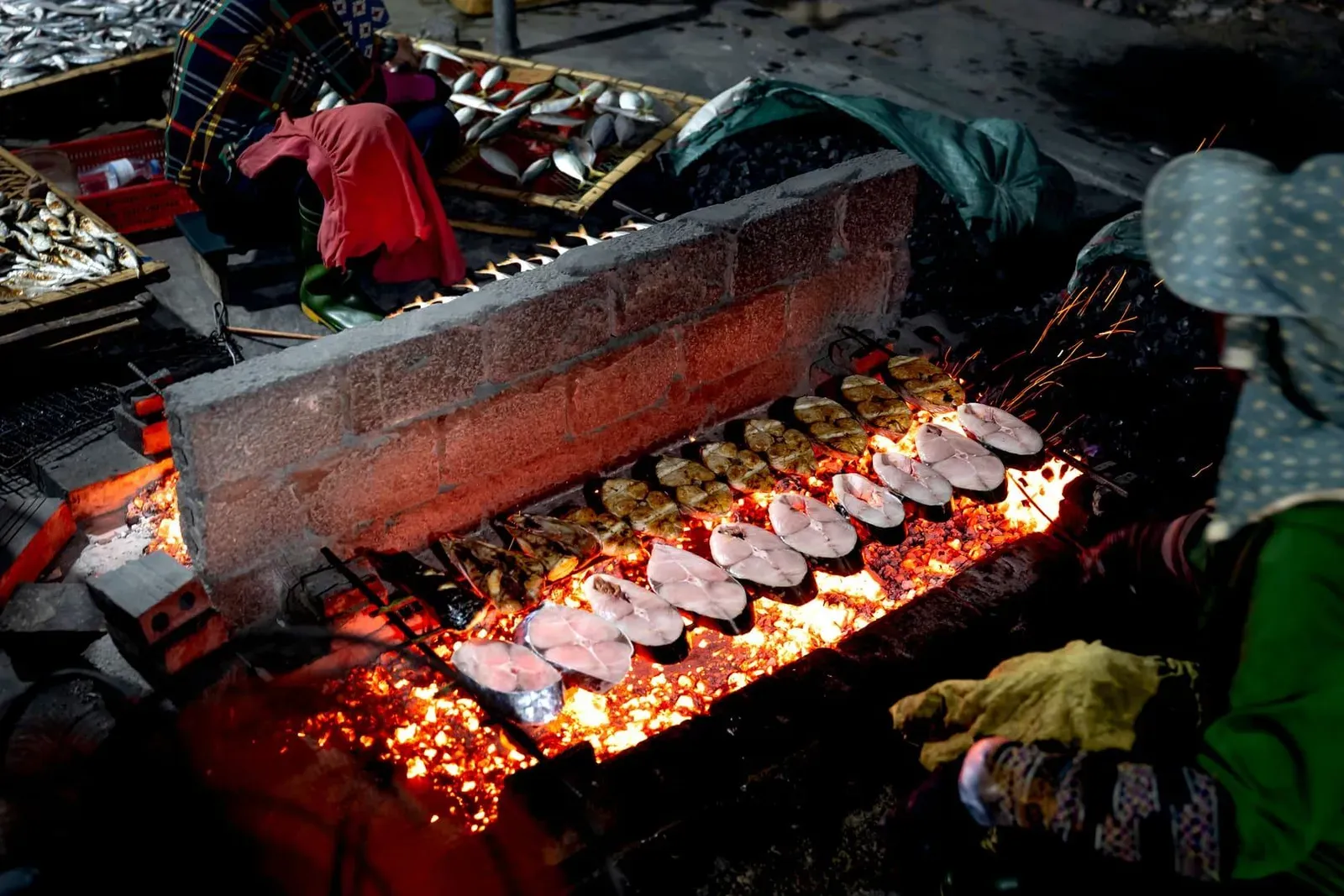 Vendor grilling fresh fish over open charcoal flames at a bustling Koh Samui street food stall at night