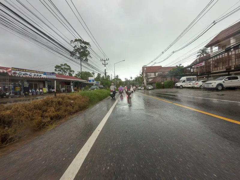 Ring Road near Lamai Beach with shopfronts and motorbike traffic on a rainy day, Koh Samui
