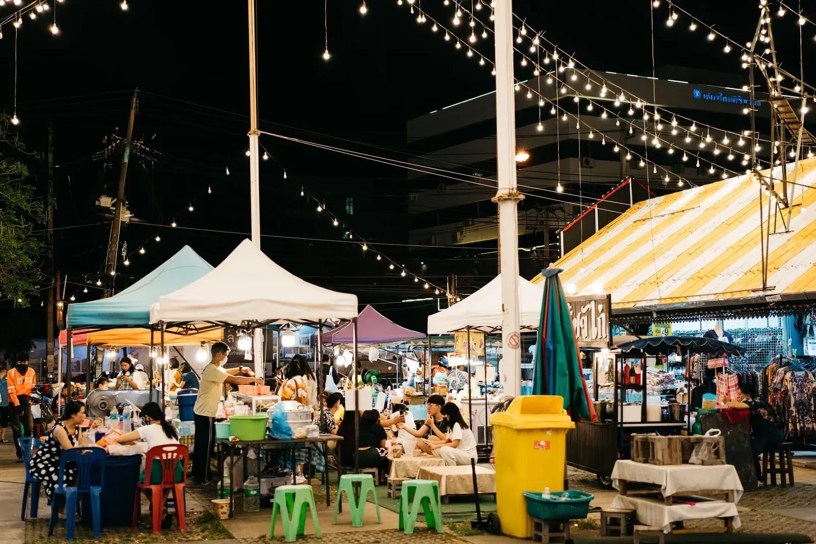 Diners sitting at plastic tables eating street food under bright market lights at night
