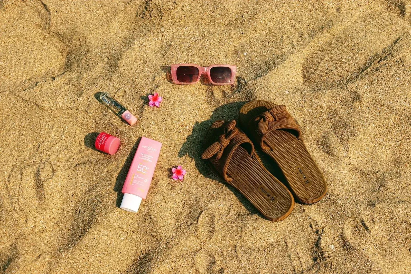Flat lay of beach packing essentials for Koh Samui including sunglasses, sandals and reef-safe sunscreen on sand