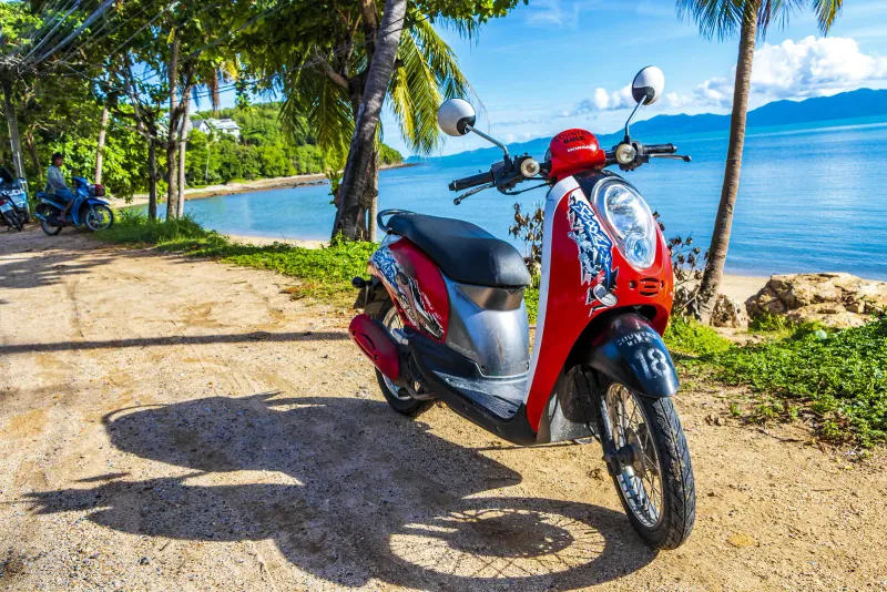 Scooter Honda rosso parcheggiato su una pista sabbiosa accanto a una baia tranquilla di Koh Samui