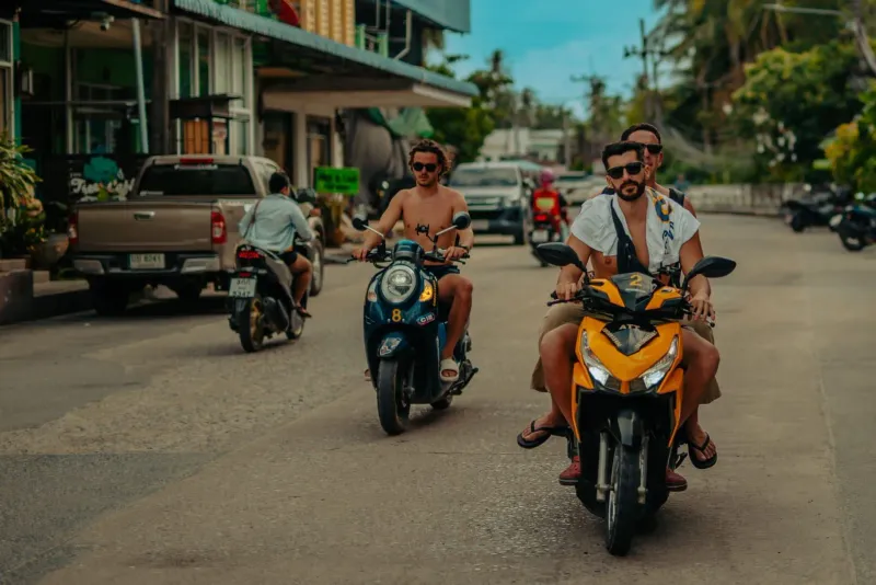 Tourists riding rental scooters without helmets down a tropical Thai street