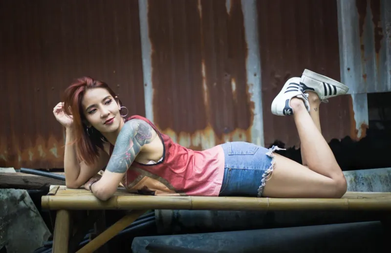 Tattooed woman poses on a wooden bench outdoors in casual tropical wear