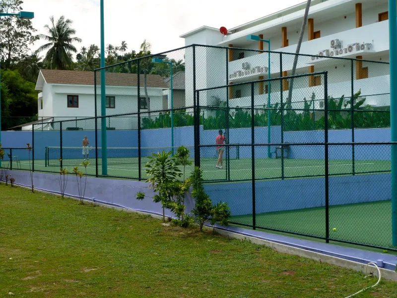 A hard court behind fence netting with a white building behind it at Koh Samui Tennis Club