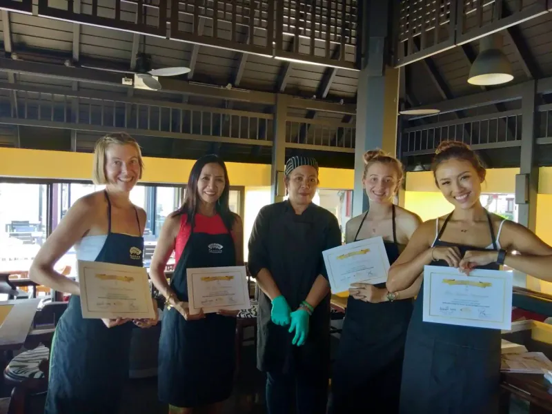 Four class participants in aprons holding certificates in an open-air cooking hall