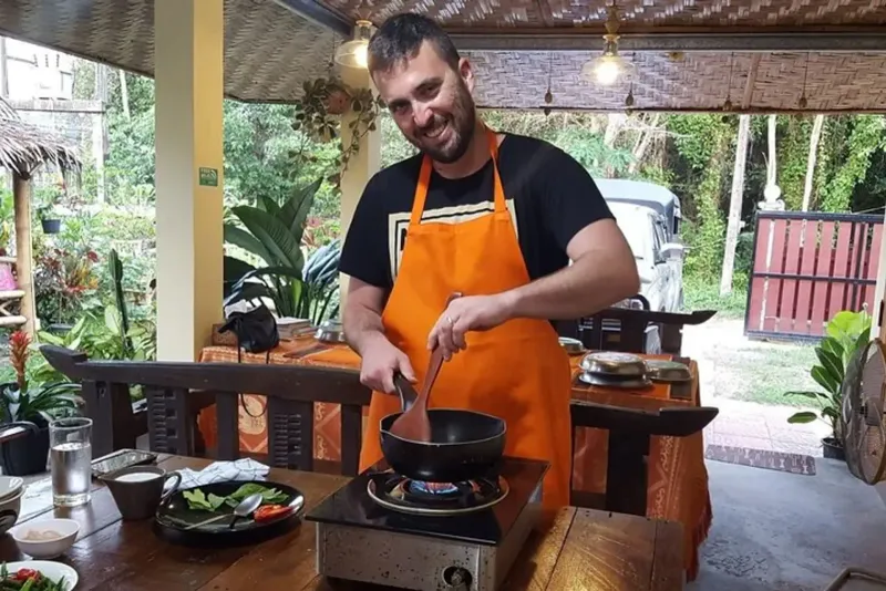 A cook in an orange apron stirring a wok in an open-air Thai cooking class kitchen