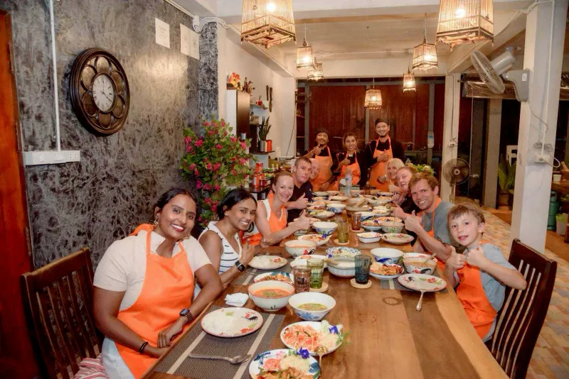 A group of cooking class guests in orange aprons around a long table of finished Thai dishes