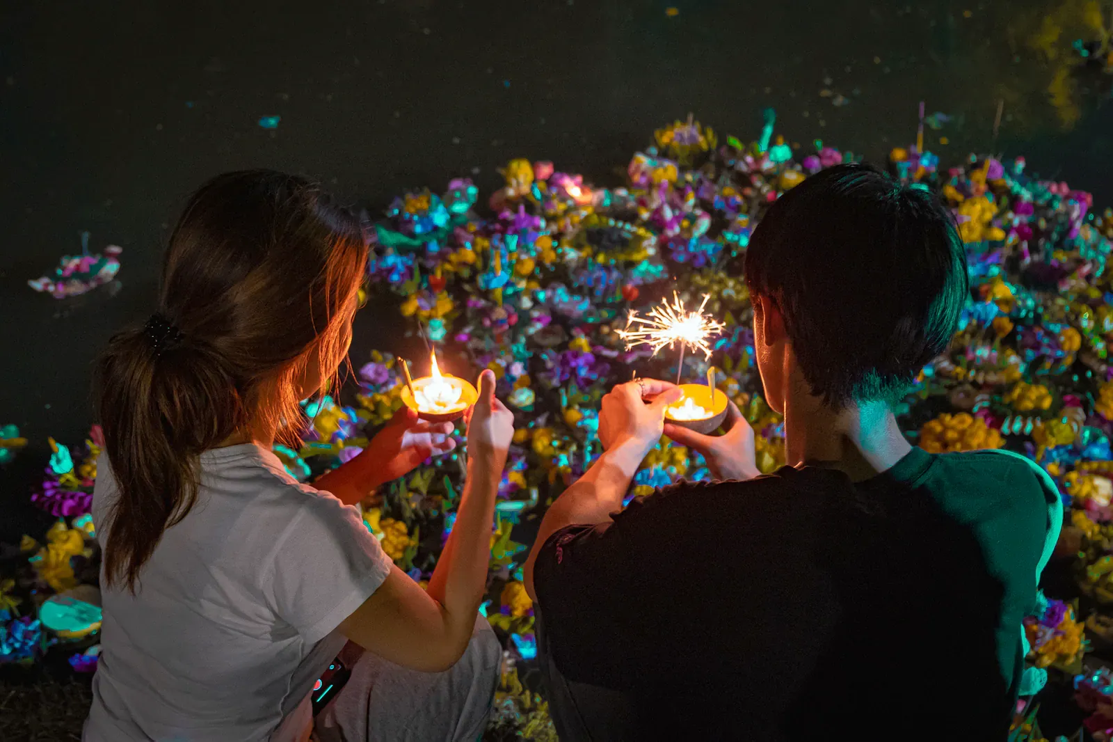 Two people holding candles and sparklers over a pile of flower krathong floats at a Loy Krathong celebration