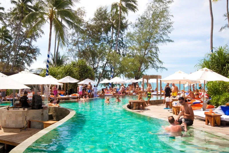 Guests in and around the curved pool at Nikki Beach Koh Samui, with white umbrellas, daybeds and the sea beyond