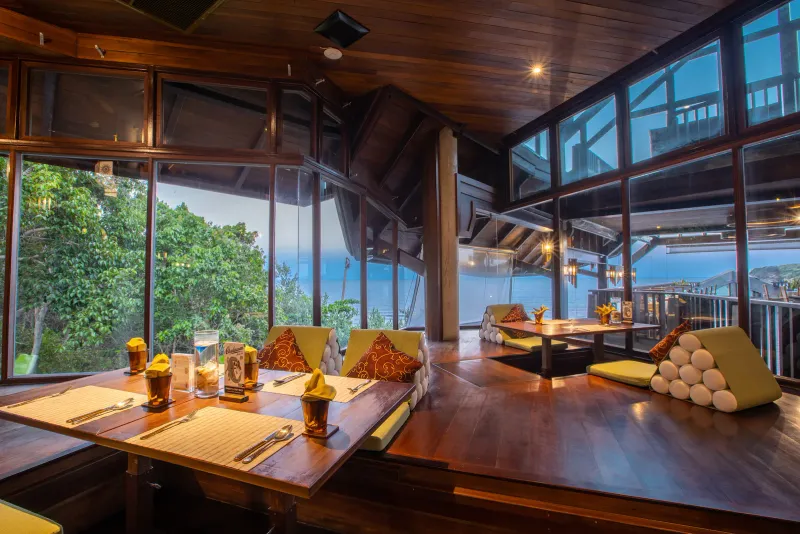 Salle à manger du Nora Buri Resort avec tables basses de style thaï et baies vitrées sur la baie