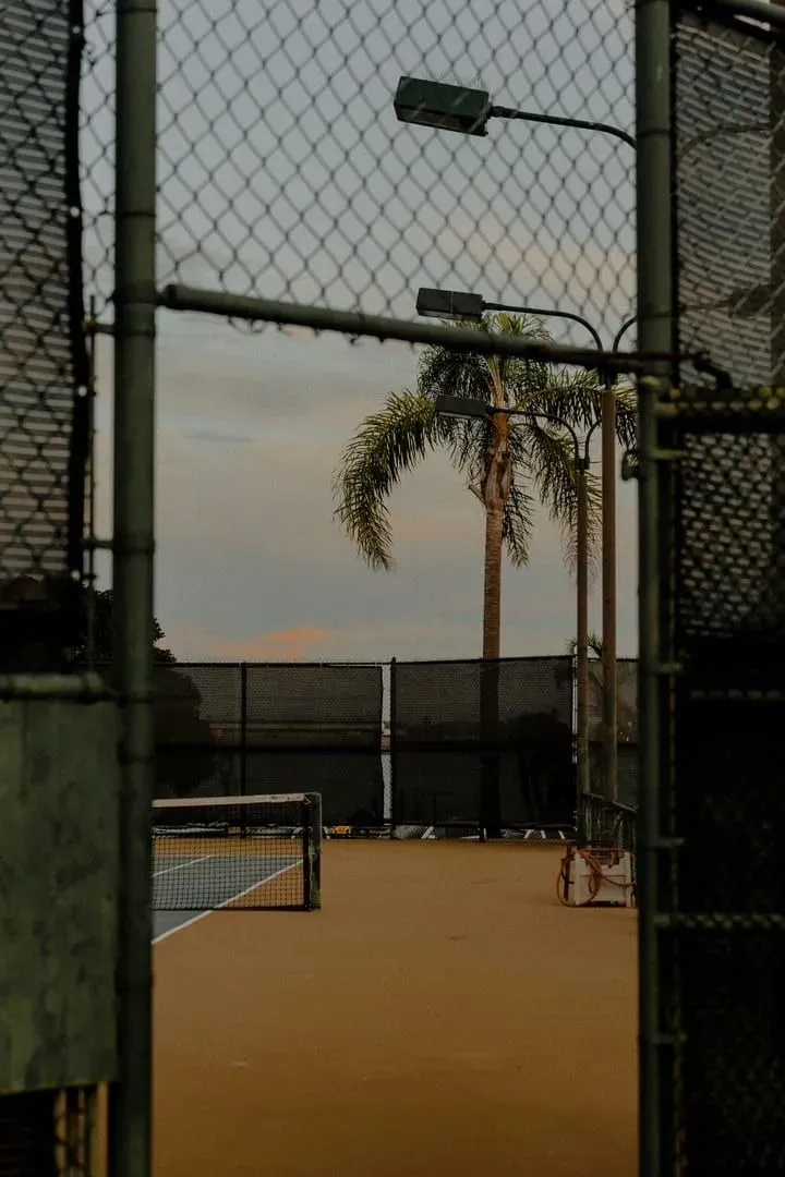 Outdoor tennis court behind a chain-link fence with palm trees at sunset, the daylight-only setup many resort courts use