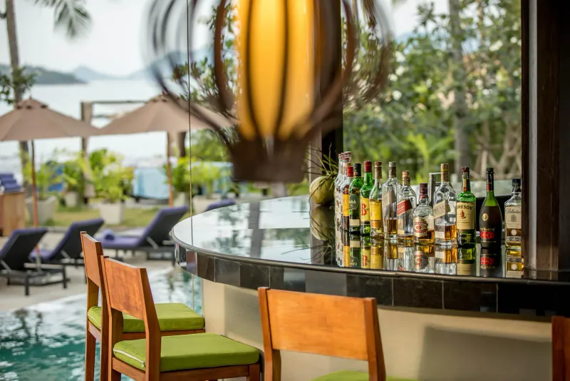 Bar in piscina con sgabelli di legno, una vetrina di bottiglie e lettini affacciati sulla baia all'Outrigger Koh Samui
