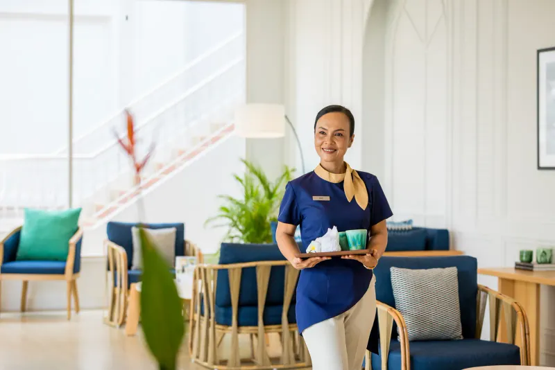 Membro del personale dell'hotel in divisa blu con un vassoio in una luminosa lounge della lobby all'Outrigger Koh Samui